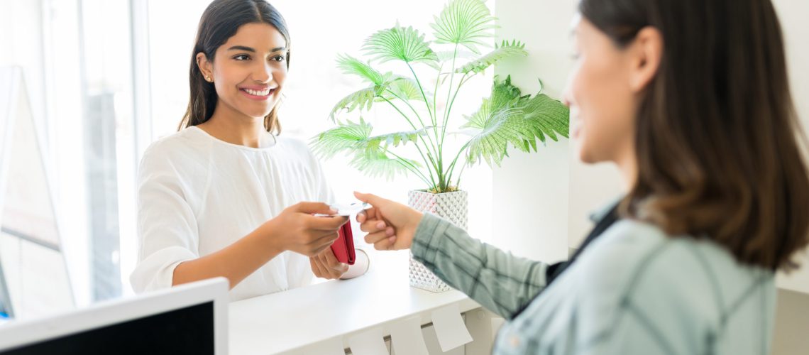 Client Paying For Beauty Treatment To Receptionist In Spa Satisfied Latin customer paying cosmetician through credit card at reception desk in beauty salon
