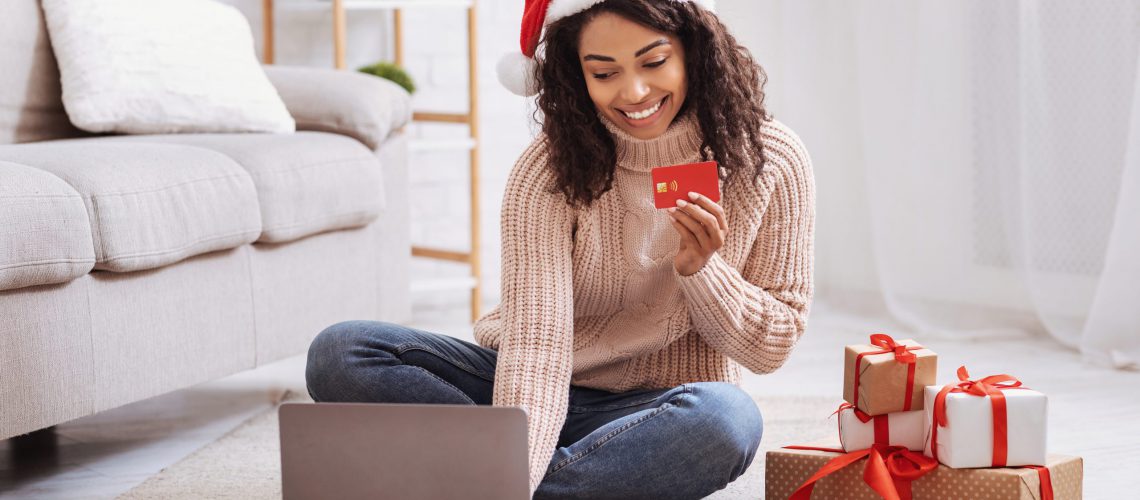 Christmas Online Shopping. Smiling black female model wearing santa hat holding red credit card and using laptop sitting in living room at home, gift boxes on the floor, ordering presents